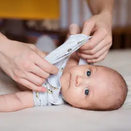 A caregiver gently dresses a baby in a light-colored onesie while the infant lies on a soft surface, showcasing a warm, homey environment.
